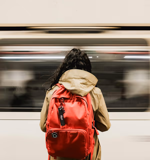 Person with long dark hair wearing a beige coat and red backpack during her travels planned on Pilot standing in front of a moving subway train.