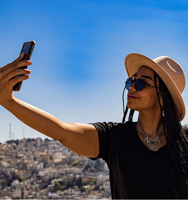Woman wearing a beige hat and sunglasses during her travels planned on Pilot taking a selfie with a cityscape in the background under a clear blue sky.