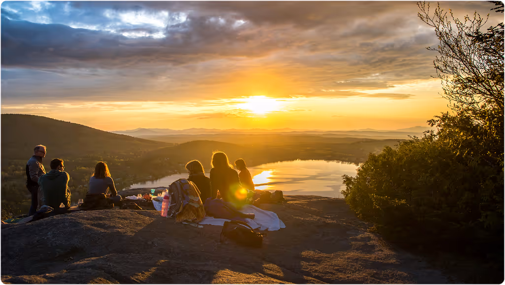 Group of people during their trip planned on Pilot. sitting on rocks watching the sunset over a lake with mountains in the background.