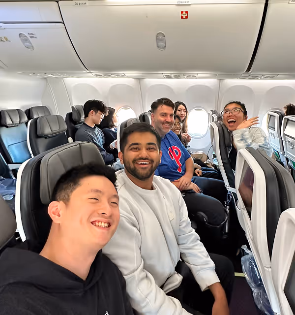 Group of six diverse people smiling and posing on an airplane during their trip planned on Pilot with rows of seats and overhead compartments visible.