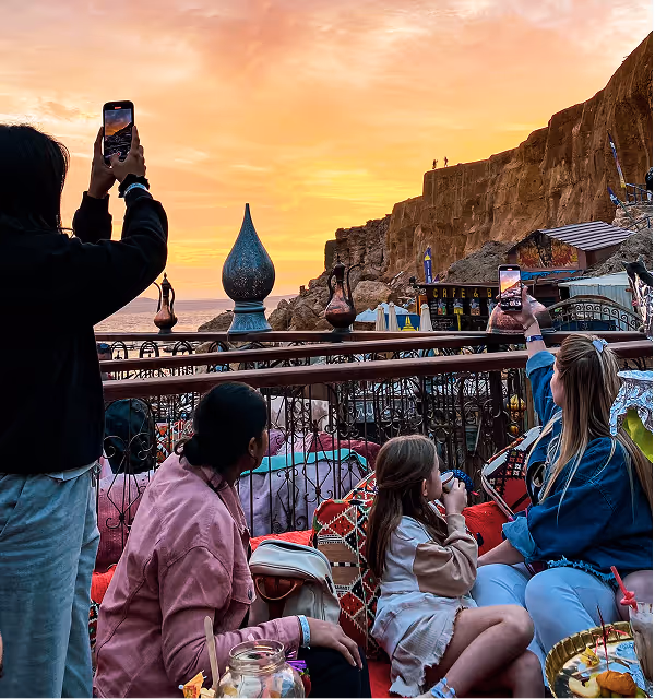Four people sitting and standing near a railing at sunset during their trip planned on Pilot with two of them taking photos of a scenic rocky coastline and orange sky.