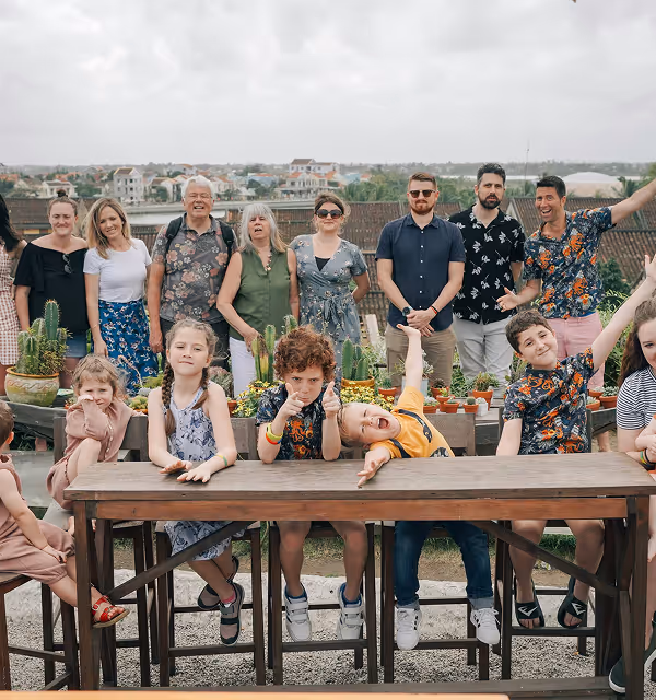 A diverse group of adults standing behind a row of children during their trip planned on Pilot sitting on stools by wooden tables outdoors with a cityscape in the background.