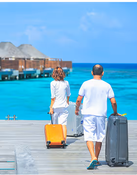 Couple in white clothing walking on a wooden pier carrying suitcases towards overwater bungalows while on a trip planned on Pilot with clear turquoise ocean and blue sky.