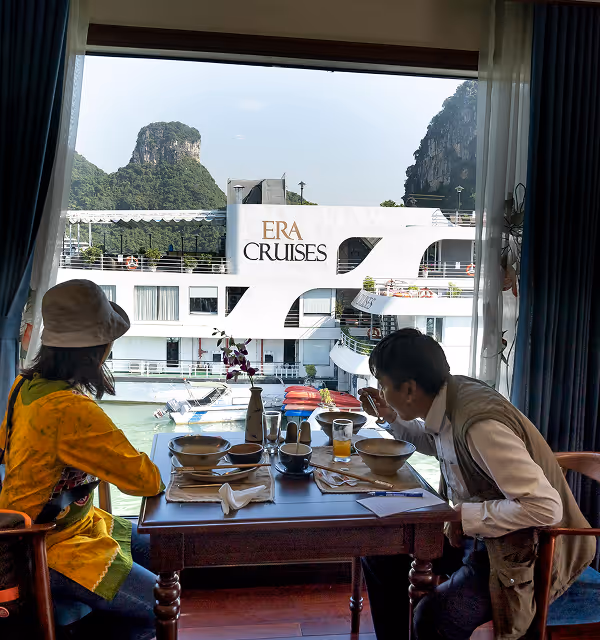 Two people while on a trip planned on Pilot dining at a table inside a room with a large window overlooking an ERA Cruises boat on the water and green limestone hills in the background.