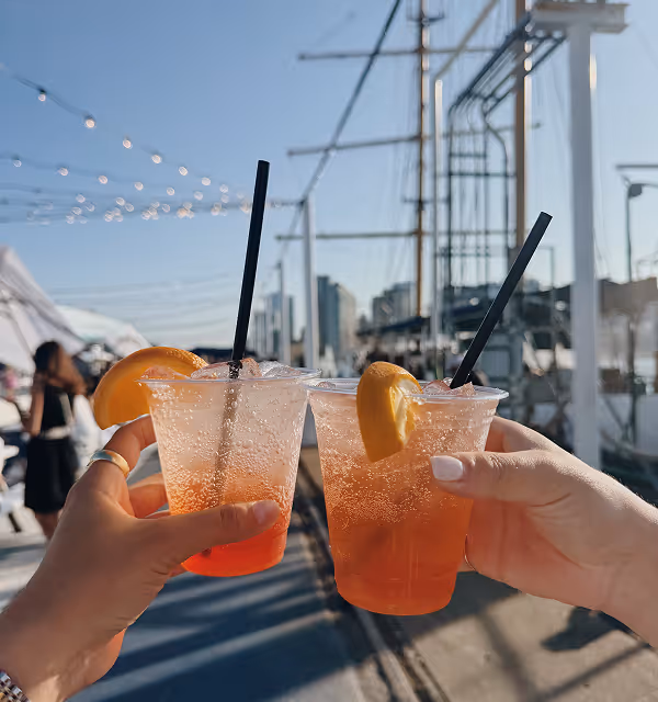 Two hands holding plastic cups with orange cocktails garnished with citrus slices, while on a trip planned on Pilot, with a marina and boats in the background.