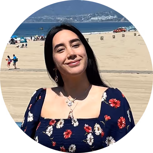 Smiling woman who works for Pilot with long dark hair wearing a floral dress at a sunny beach with ocean and distant hills.