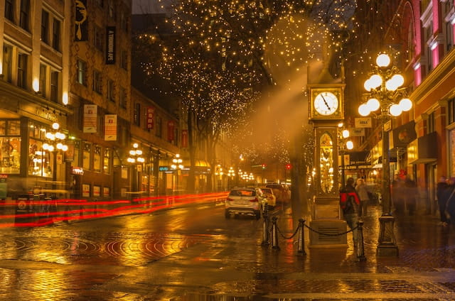 A clock tower in the street with yellow lights and car parks by the sidewalk