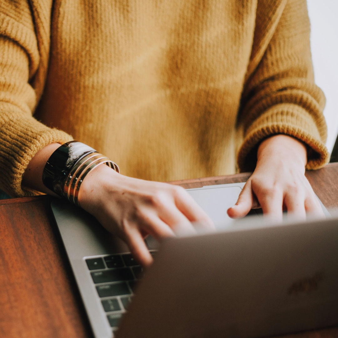 Person wearing a mustard yellow sweater typing on a laptop at a wooden table, with a silver and black bracelet on the left wrist.