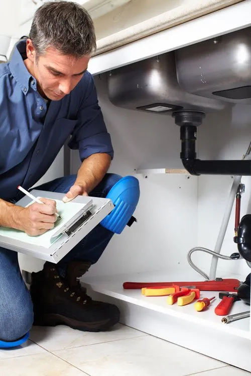 Plumber in blue uniform working under kitchen sink with tools and clipboard