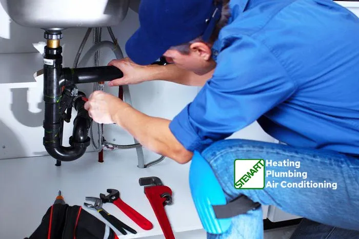 A male plumber wearing a blue cap, blue shirt, and blue jeans with blue knee pads is crouched beneath a sink, actively working on the pipes. His hands are engaged with a wrench on the black PVC piping. On the floor beneath the sink, various tools, including red pipe wrenches and a black tool bag, are neatly laid out. In the bottom right corner, a green and white logo for "STEWART Heating Plumbing Air Conditioning" is displayed.