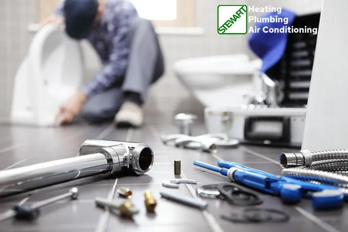 A plumber, blurred in the background, is working on a toilet, with his back partially to the camera. In the foreground, various plumbing tools and parts, including chrome pipes, a blue wrench, and fittings, are scattered across a tiled floor. In the upper right corner, a green and white logo for "STEWART Heating Plumbing Air Conditioning" is displayed.