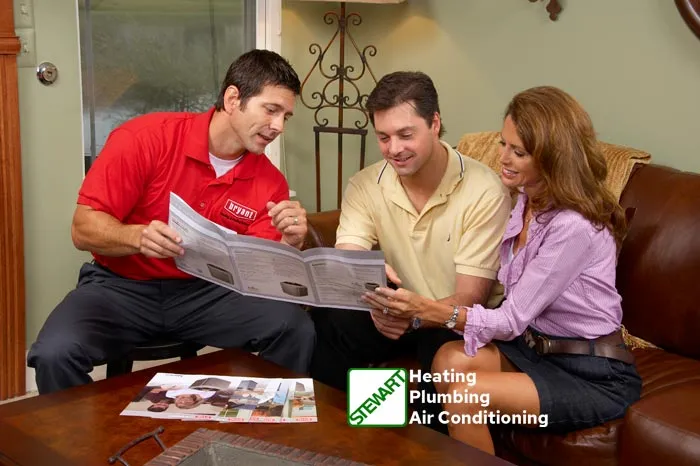 A horizontal image shows three people in a living room setting, facing each other. On the left, a man in a red "Bryant" uniform shirt is holding an open brochure and gesturing towards it. In the center, another man in a yellow polo shirt is looking at the brochure, smiling. On the right, a woman with long, curly brown hair, wearing a purple striped shirt and dark skirt, is also looking at the brochure and smiling. They appear to be seated around a coffee table, on which another brochure is visible. In the bottom right corner, a green and white logo for "STEWART Heating Plumbing Air Conditioning" is displayed.
