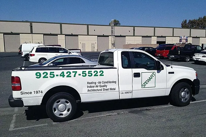 
A white pickup truck with the "STEWART" logo on its side is parked in an outdoor lot. The truck is adorned with green lettering that includes a phone number: "925-427-5522," and a list of services: "Heating, Air Conditioning, Indoor Air Quality, Architectural Sheet Metal." Below the phone number, the text "Since 1974" is visible, and on the lower right, "Lic. #290747" is displayed. A silver car is parked in the truck's bed. In the background, there's a large building with multiple garage doors and other vehicles parked in the lot.
