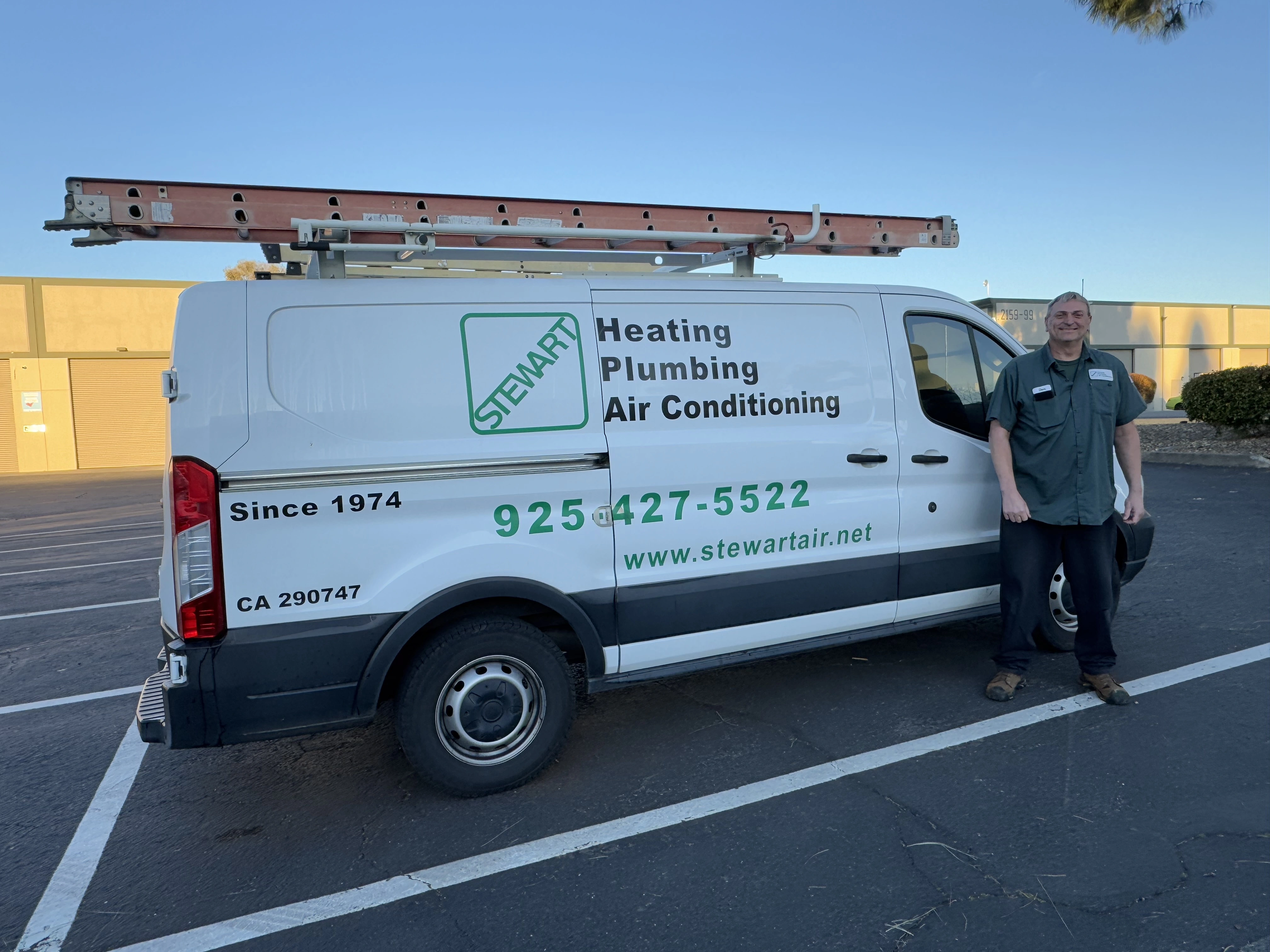 A man in a green uniform shirt and dark pants stands next to a white service van. The van has the logo "STEWART Heating Plumbing Air Conditioning" in green text on its side. Below that, in black text, are "Since 1974" and "CA 290747". A phone number "925 427-5522" and a website "www.stewartair.net" are also displayed on the van in green. A ladder is secured to the roof rack of the van. The background shows a building and what appears to be a parking lot.