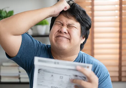 A man with dark, disheveled hair, wearing a blue t-shirt, is shown in a close-up shot, appearing frustrated or distressed. He is holding a blurred document, possibly a bill or statement, with his left hand while his right hand is scratching his head. His eyes are squinted shut and his face is contorted, conveying strong negative emotion. In the background, there's a hint of indoor setting with a blurred plant and window blinds.