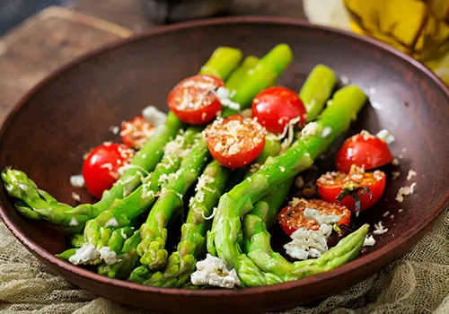 A close-up, high-angle shot of a vibrant green asparagus and cherry tomato dish served in a rustic brown bowl. Several stalks of blanched or steamed asparagus are arranged in the bowl, topped with bright red cherry tomatoes, some whole and some halved. The dish is garnished with crumbled white cheese or feta and possibly some herbs. In the blurred background, a glass bottle with olive oil is partially visible.