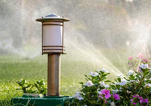 A garden scene featuring a decorative outdoor lamp post in the foreground. The lamp post has a cylindrical white light fixture enclosed in a dark, possibly metal, frame with horizontal bars, topped with a cap. It stands on a green base. To the right of the lamp post, there are green bushes with white and pink flowers in bloom. In the background, a lawn sprinkler is actively spraying water, creating a misty effect and arcs of water visible against the green grass. The sunlight illuminates the water spray.