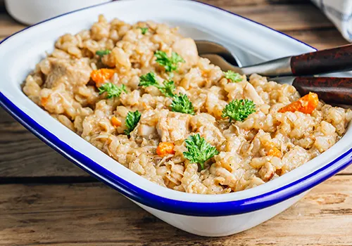 A close-up, elevated shot shows a white rectangular dish with a blue rim filled with a hearty-looking stew. The stew consists primarily of grains, possibly barley or rice, mixed with pieces of meat or poultry and diced orange vegetables, likely carrots. It is garnished with several small sprigs of fresh parsley. Two serving spoons with dark wooden handles are partially visible in the back of the dish. The dish rests on a rustic wooden surface.