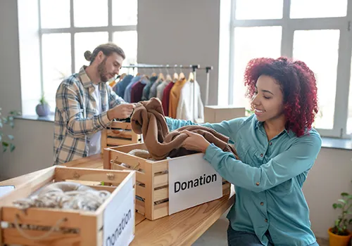 A smiling woman with reddish-brown curly hair, wearing a teal button-up shirt, is placing folded clothing into a wooden crate labeled "Donation" on a light-colored wooden table. Behind her, to the left, a man with a beard and a plaid shirt is also handling clothes, with a clothing rack full of garments visible in the background. Two large windows are visible in the background, providing natural light.