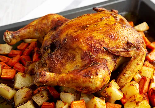 A whole roasted chicken, golden brown and glistening, is presented in a dark rectangular baking pan. It is surrounded by a generous bed of roasted vegetables, including diced carrots and potatoes that are slightly caramelized and tender. The chicken is the centerpiece, with its skin appearing crispy and seasoned.