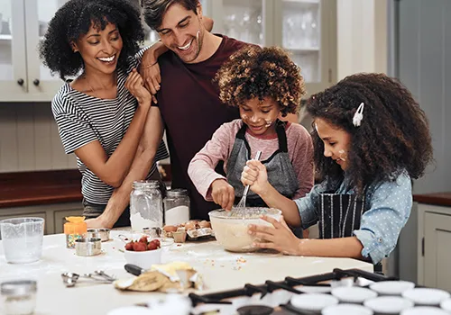 
A family of four is gathered around a kitchen counter, engaged in baking. From left to right, a woman with dark curly hair in a striped shirt stands smiling with her arm around a man in a maroon shirt, who is also smiling. In front of them, two children with curly hair are actively involved in the baking process. One child, wearing a dark apron, is mixing batter in a large bowl, while the other child, also in an apron, watches with a smile. Both children have flour or batter smudges on their faces. 