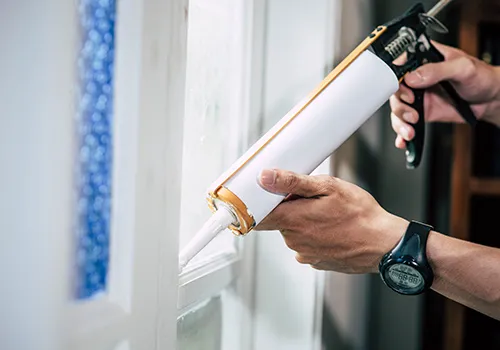 A close-up shot focuses on a person's hands operating a white and yellow caulk gun. The person is applying a bead of white caulk along the frame of a window or similar fixture. The hands are prominent in the foreground, with the caulk gun held firmly. The background is blurred, showing a windowpane with a blue patterned design on the left, and a darker, less defined area on the right, possibly an interior space.