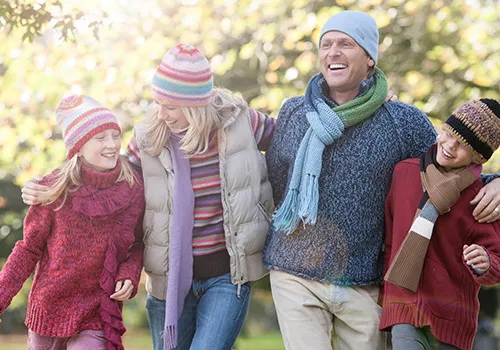 
A family of four, appearing to be two adults and two children, stands outdoors in bright sunlight, all dressed in warm winter clothing. They are positioned from left to right: a young girl in a red sweater and striped hat; a woman in a striped shirt, puffer vest, and long purple scarf; a man in a blue knit sweater, light blue beanie, and a green and blue scarf; and another young child in a red sweater, knit hat, and brown scarf.