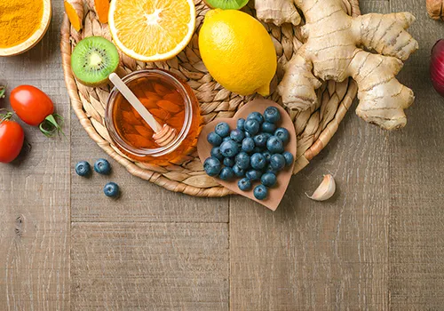 
A flat lay shot showcases various healthy foods arranged on a rustic wooden surface. In the upper half of the image, a woven, circular placemat holds a jar of honey with a wooden dipper, half a kiwi, a sliced orange, a whole lemon, and several pieces of ginger. To the left of the placemat, a small wooden bowl contains a pile of turmeric powder, and two red cherry tomatoes are visible.