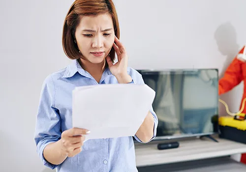 A medium close-up shot of a light-skinned woman with short, auburn hair, dressed in a blue button-up shirt. Her brow is furrowed, and her right hand is on her temple, indicating concern or confusion, as she holds and looks at a stack of white papers in her left hand. A television screen and what appears to be a tool box are visible in the background.