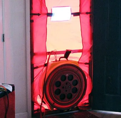 A bright red fabric frame, secured in a doorway, houses a large, yellow, circular fan, which is centered in the lower half of the frame. The fan's opening is black with several circular cutouts. Wires from the fan lead to the top of the frame.