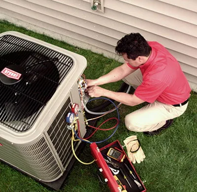 A high-angle shot captures a male technician, wearing a red polo shirt and khaki pants, kneeling on a green lawn while performing maintenance on an outdoor Bryant air conditioning unit. He is connecting blue and red hoses from a set of gauges to the unit's service ports. An open red toolbox is on the grass, containing various tools including a yellow multimetre. A pair of work gloves is also visible beside the toolbox.