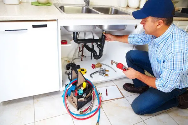 A plumber in a blue cap is kneeling and working on the pipes under a kitchen sink, with a tool bag and tools scattered on the tiled floor around him.