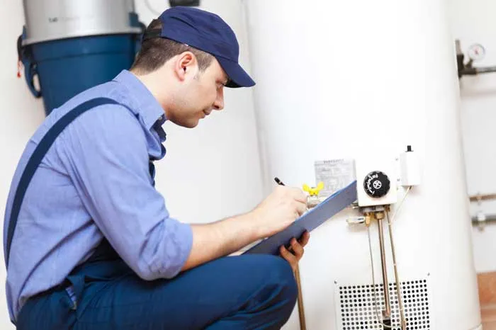 A technician in a blue cap and overalls is crouching and looking at a water heater, holding a clipboard and pen to take notes.