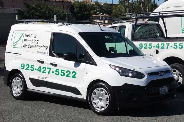 A white commercial van with the "Stewart Heating Plumbing Air Conditioning" logo and contact information in green text is parked on a paved lot on a sunny day.