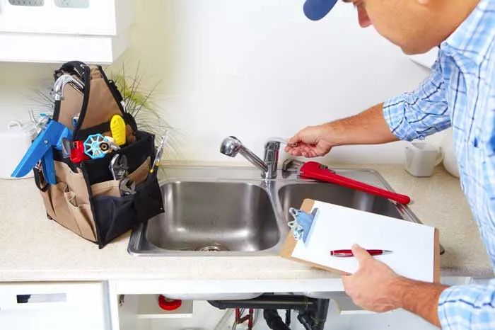 A man wearing a blue cap and plaid shirt is seen from the waist up, adjusting a silver faucet over a double-basin kitchen sink, with a tan tool bag filled with various plumbing tools on the counter to his left. He holds a clipboard with paper and a pen in his left hand.