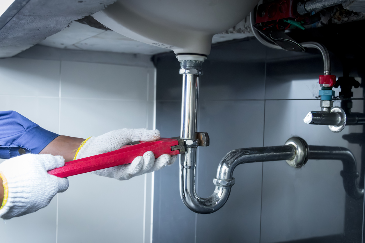 A close-up shot shows a plumber's gloved hands using a red wrench to work on the p-trap and pipes underneath a white sink.