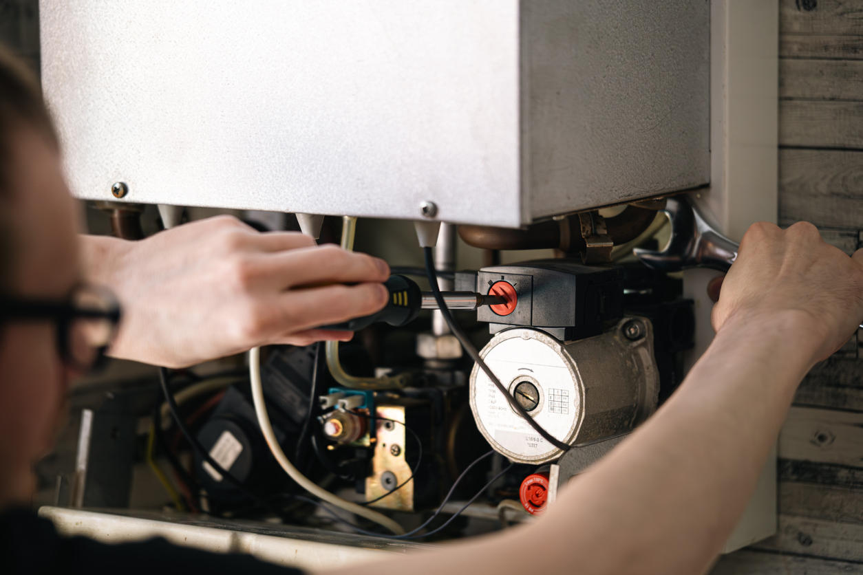 A man wearing glasses is working on a tankless water heater mounted on a gray tiled wall, using a wrench and surrounded by tools and copper fittings on the floor.
