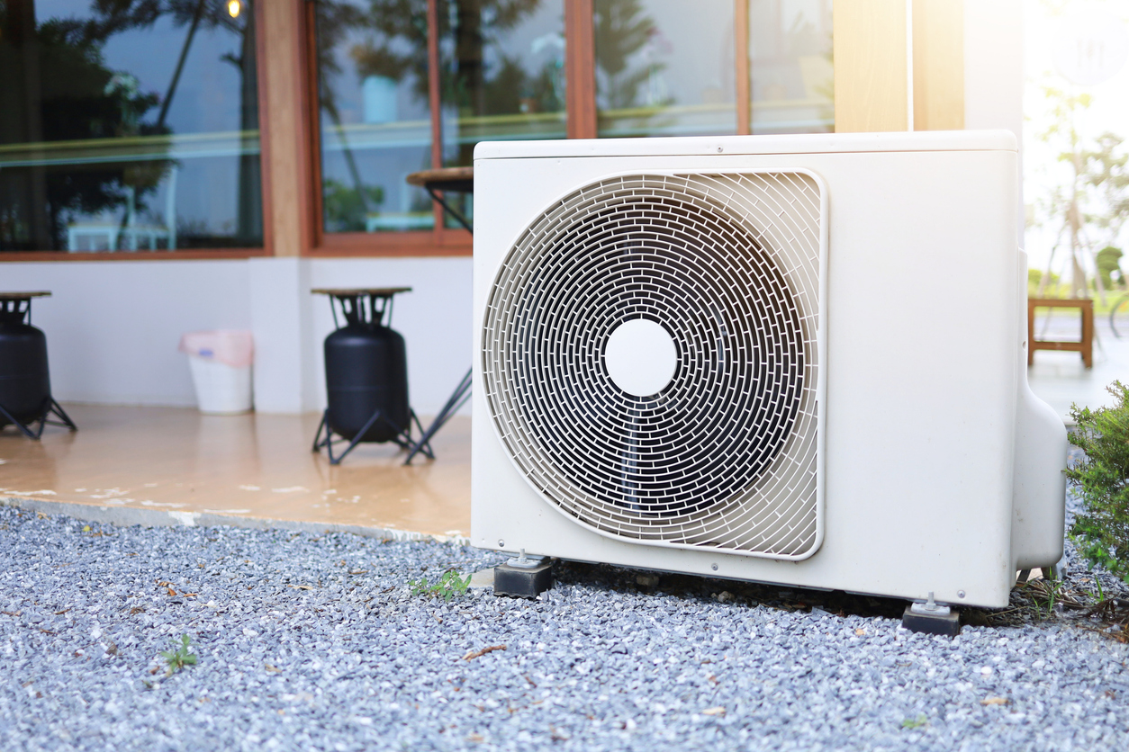 A heat pump is installed on a bed of gravel next to a light brown building with large glass windows, casting shadows on the ground under a bright sun.
