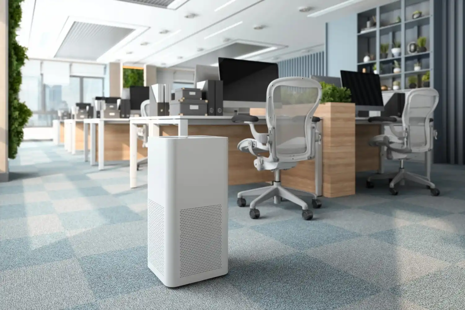  A modern, minimalist white air purifier with a perforated front is positioned in the foreground of a brightly lit, open-plan office space with numerous desks and chairs.