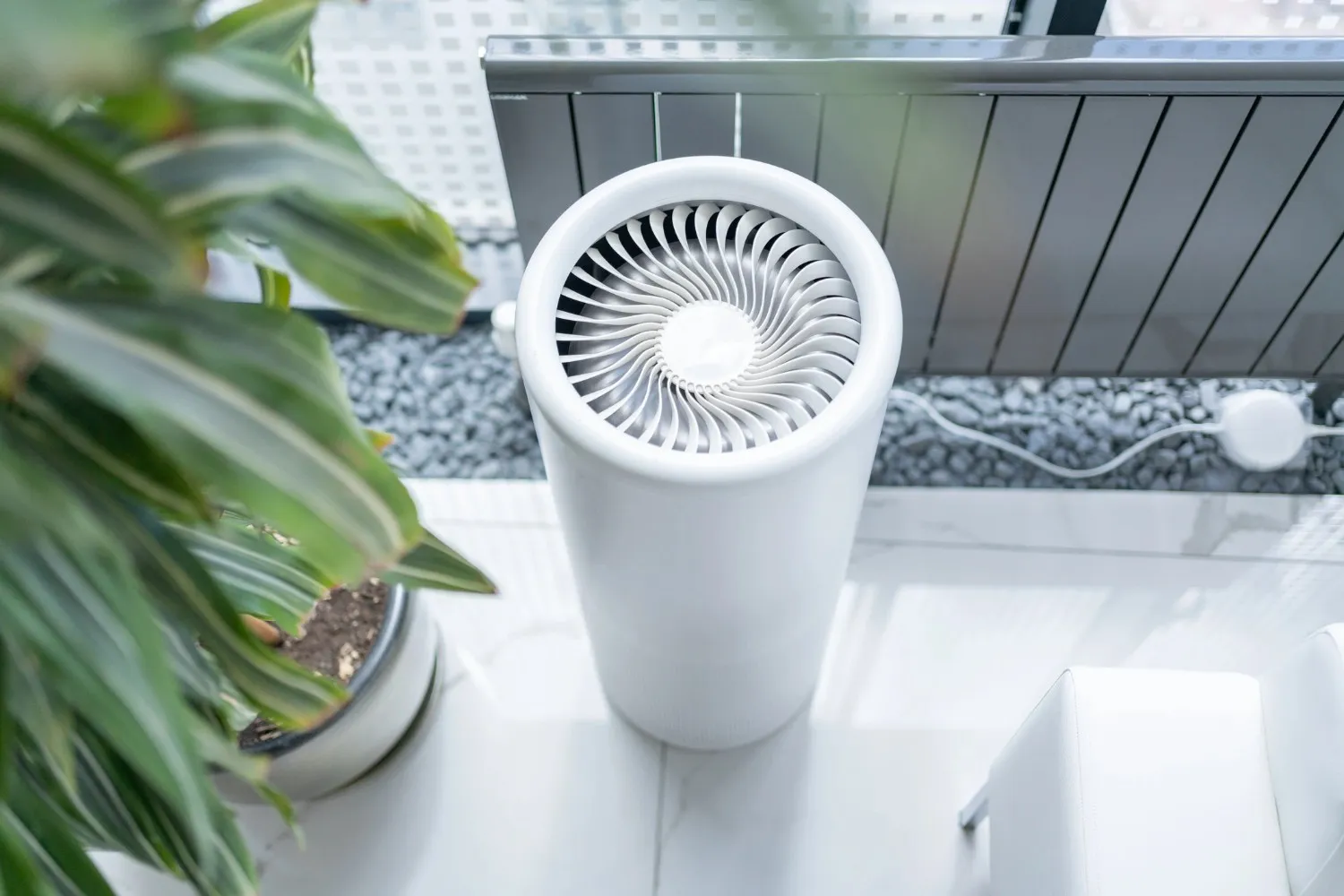 An overhead view of a white cylindrical air purifier with a spiraled top fan is positioned on a white tiled floor next to a green plant, with a blurred outdoor area in the background.