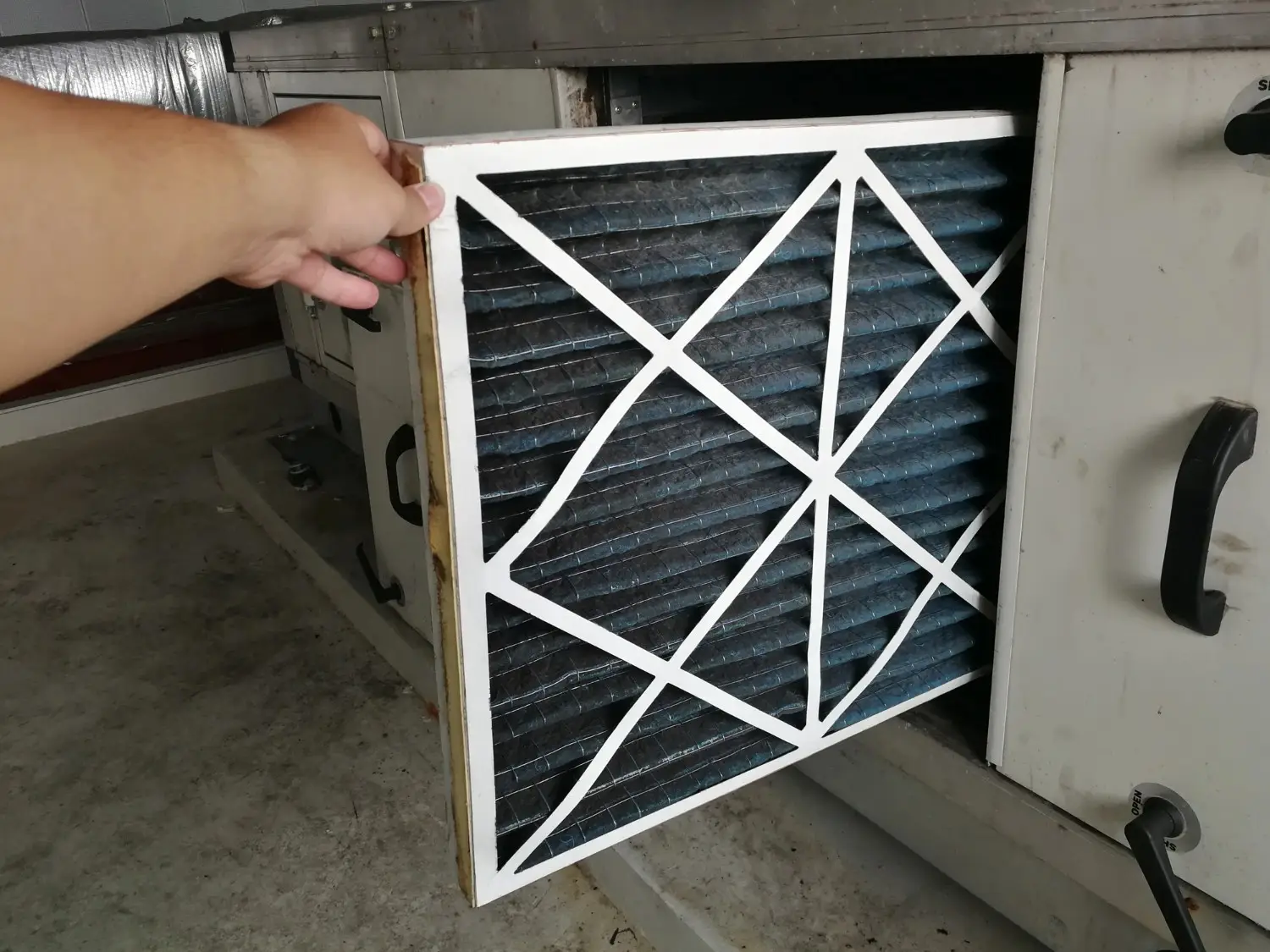  A hand is pulling out a dirty, pleated air filter with white reinforcement strips from a large, off-white HVAC unit.