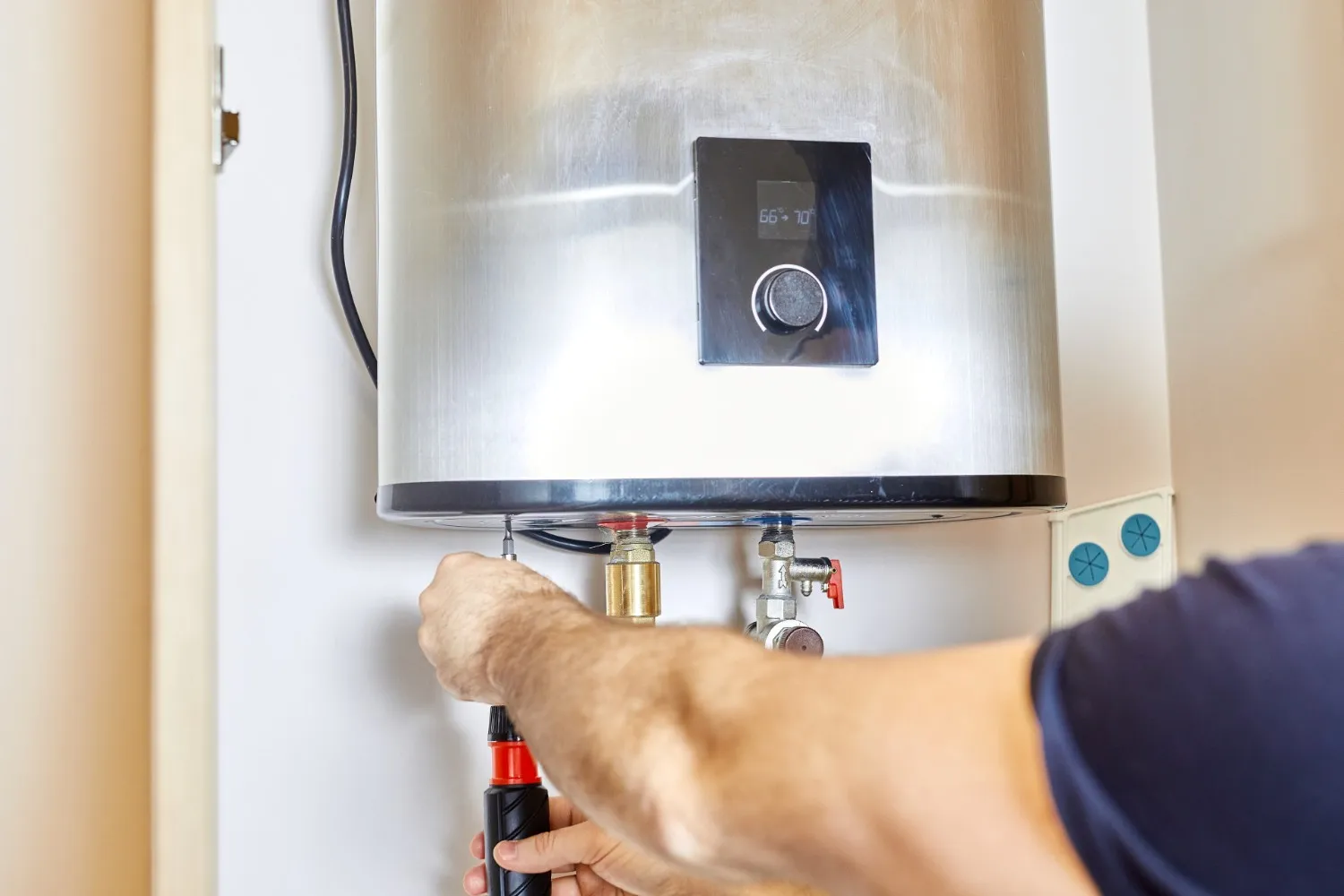 A close-up shot shows a person's hands, wearing a dark blue shirt, working on the bottom of a shiny, metallic water heater. The water heater has a black digital display with the numbers "66° 70°" visible, and a circular knob below it. The person is holding a tool with a red and black handle, seemingly adjusting a pipe or valve connected to the water heater.