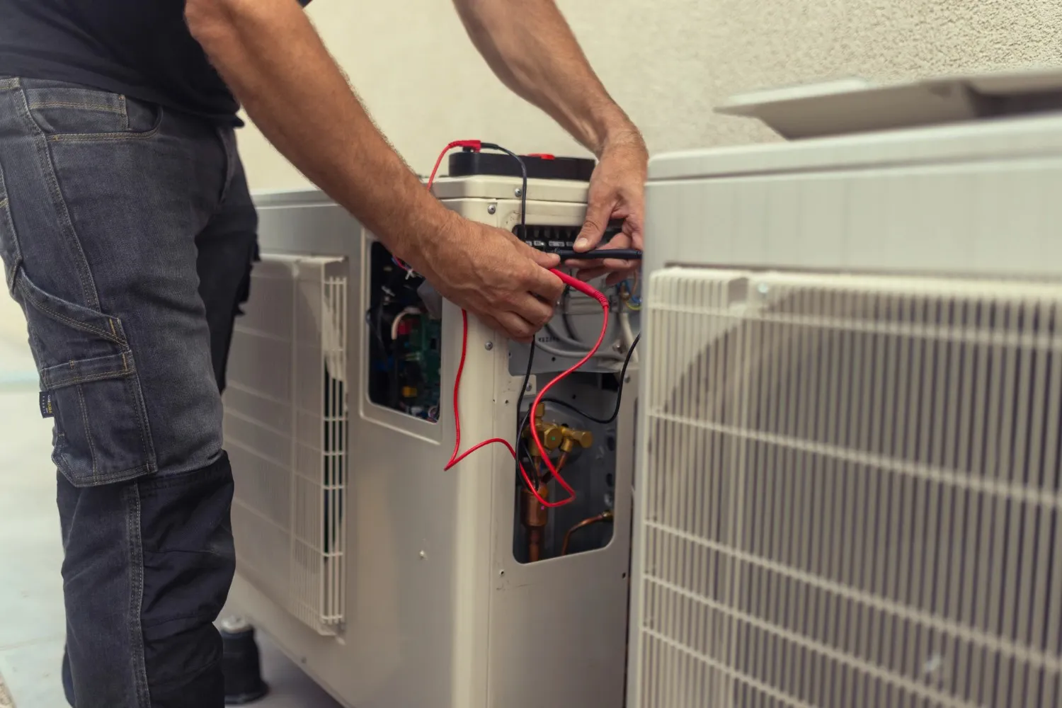 A male technician, wearing dark work pants and a dark t-shirt, is actively performing diagnostics or repair on an outdoor HVAC unit. He is using a multimeter, holding its red and black probes to test electrical connections within the open panel of the unit, revealing wires and circuitry. Another similar outdoor unit is partially visible to the right.
