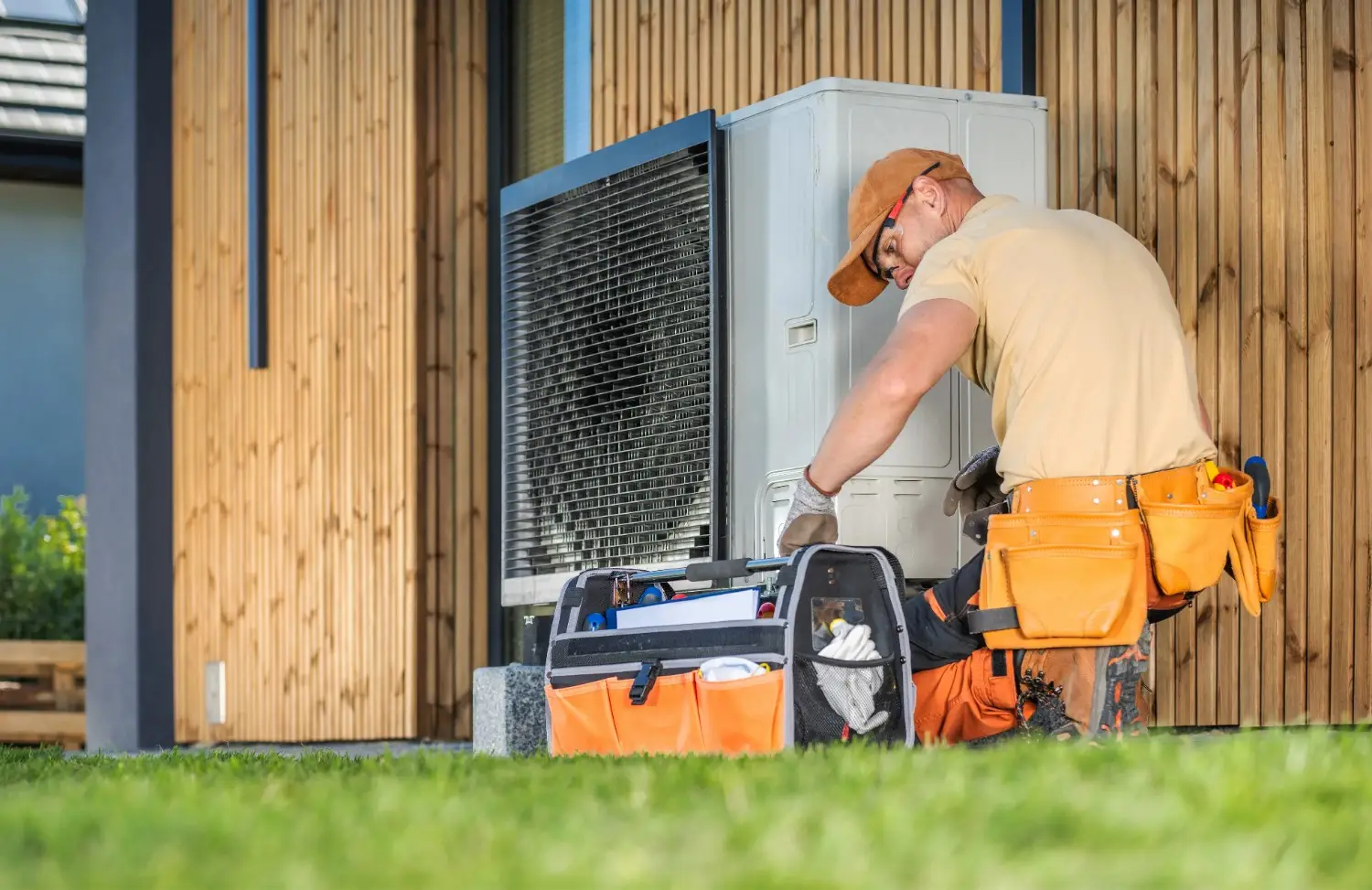 A male technician, wearing a tan t-shirt, orange shorts over work pants, an orange baseball cap, and work gloves, is actively working on an outdoor heat pump or air conditioning unit. He is bending over the unit, which is white and black, and has a large grill on its side. To his left, an orange and grey open toolbox filled with tools sits on the grass.