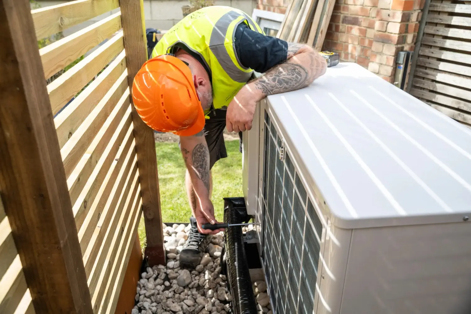 A male technician wearing a bright yellow safety vest, an orange hard hat, and dark work clothes is crouching and actively working on the side of a white outdoor heat pump unit. He is using a wrench or similar tool to tighten or loosen something near the bottom of the unit. His forearms are visible, showing tattoos. The heat pump is placed on a bed of light-colored pebbles next to a wooden slatted fence.