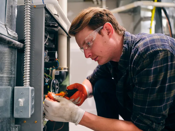 A focused young HVAC technician wearing safety glasses and work gloves is using a tool to work on the electrical wiring inside the open panel of a furnace or air handler unit.
