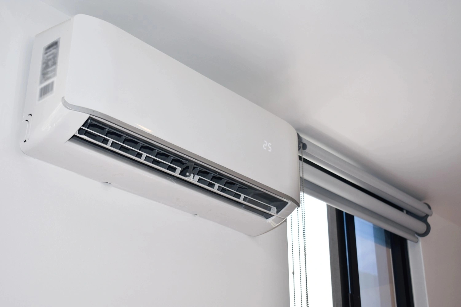 A horizontal, rectangular, white ductless AC unit is mounted high on a white wall, displaying a temperature of 25 degrees Celsius on its digital screen.