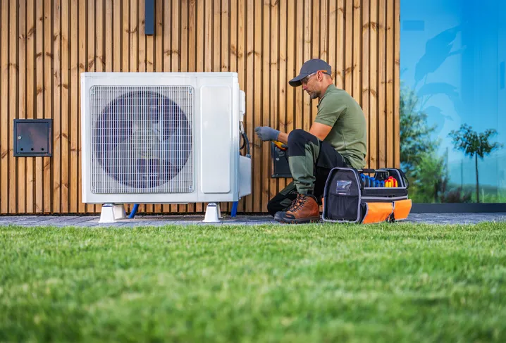 HVAC technician servicing outdoor AC unit beside wooden building with tools in grassy area