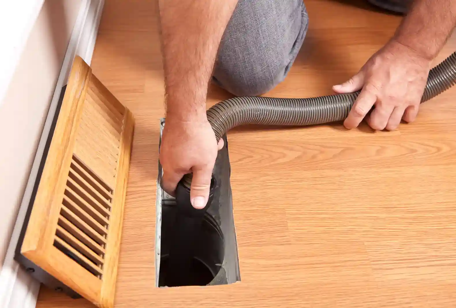 A person is kneeling on a wooden floor, using a gray vacuum hose to clean a rectangular floor vent, with the wooden cover removed and placed beside it.