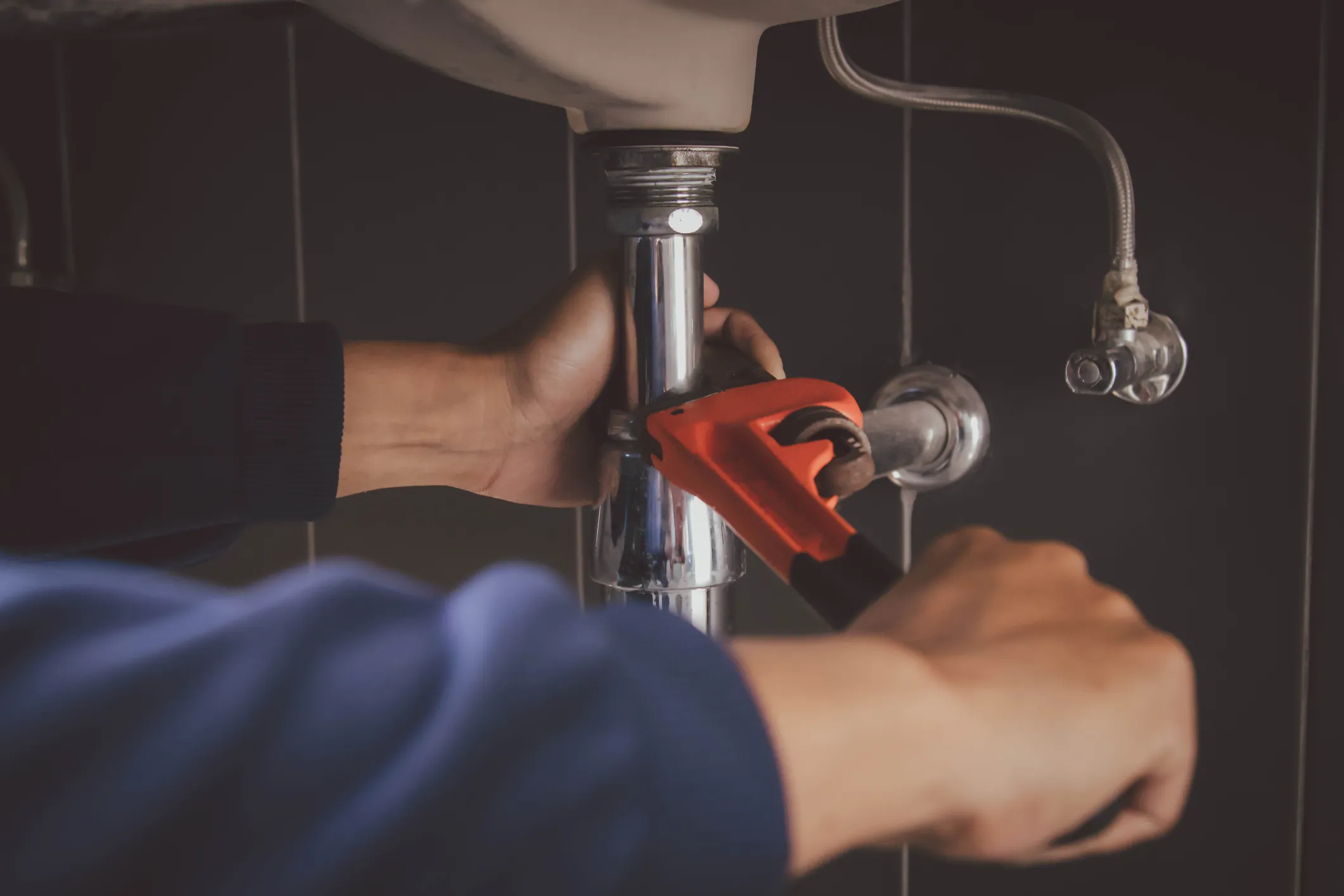  A plumber's hands are shown, one wearing a blue long-sleeved shirt and the other a black short-sleeved shirt, working on a chrome P-trap pipe beneath a white sink basin with an orange wrench.